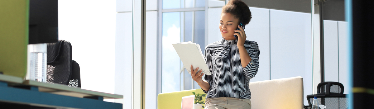 Woman standing in office talking on cellphone  while looking at papers in her hand