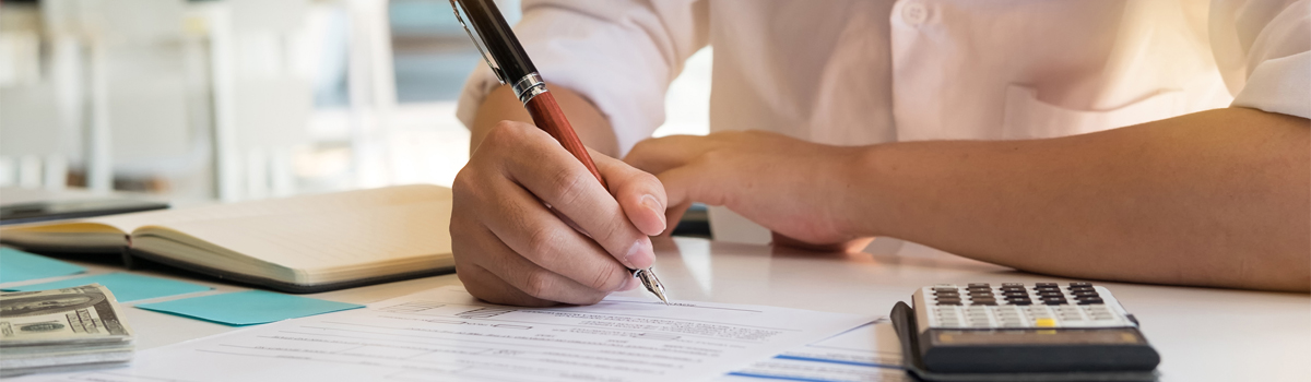 Close up of hand signing document at desk with calculator, money and notebook