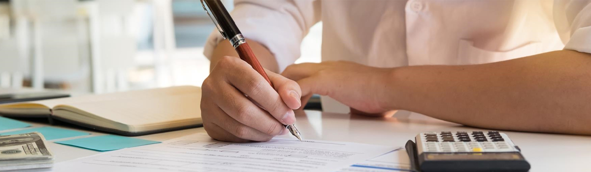 Close up of hand signing document at desk with calculator, money and notebook