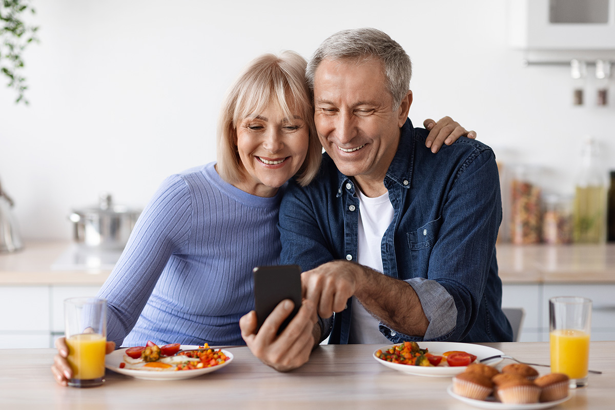 Couple sitting at breakfast table looking at smartphone smiling