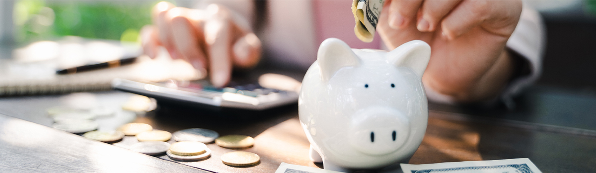 A person putting a coin into a piggy bank while using a calculator.