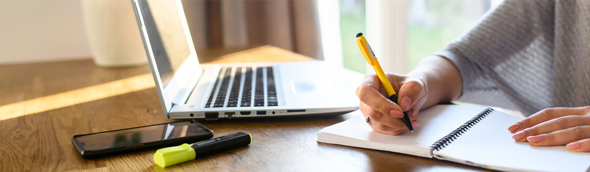 female writing in notebook at a desk with laptop and smartphone.